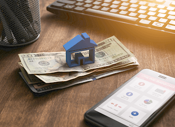 Blue house plastic piece on top of a pile of cash beside a phone with a banking app open