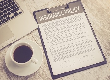 Paper that reads insurance policy on a clipboard beside a cup of coffee and laptop